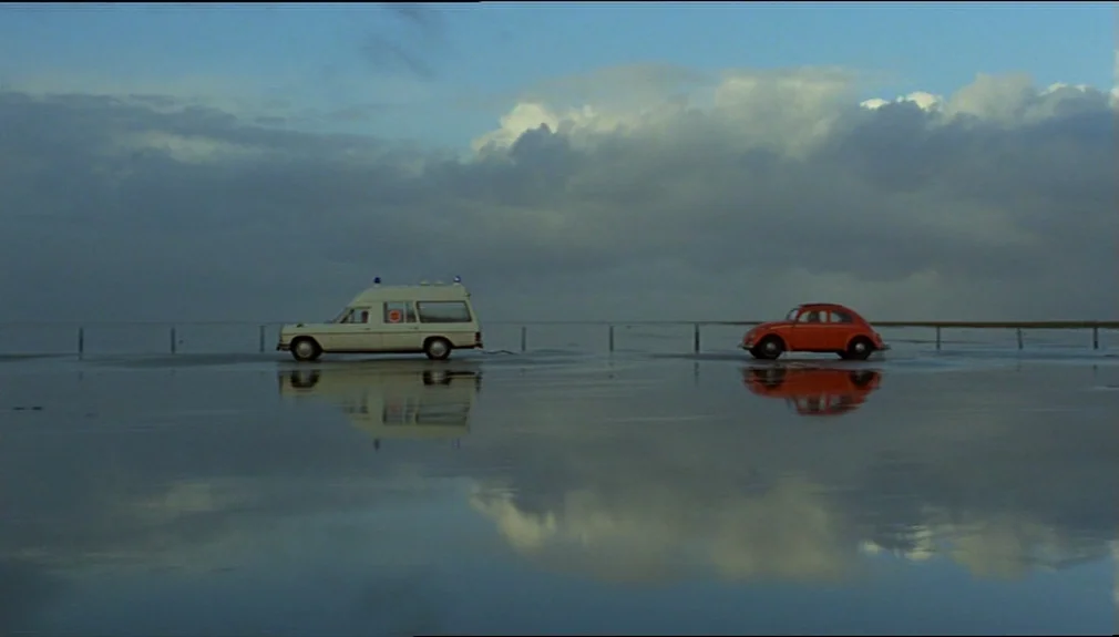 The American Friend (1977) movie still: wide — A wide, flat tidal area with a wet surface reflecting the cloudy sky. An ambulance is towing a small…
