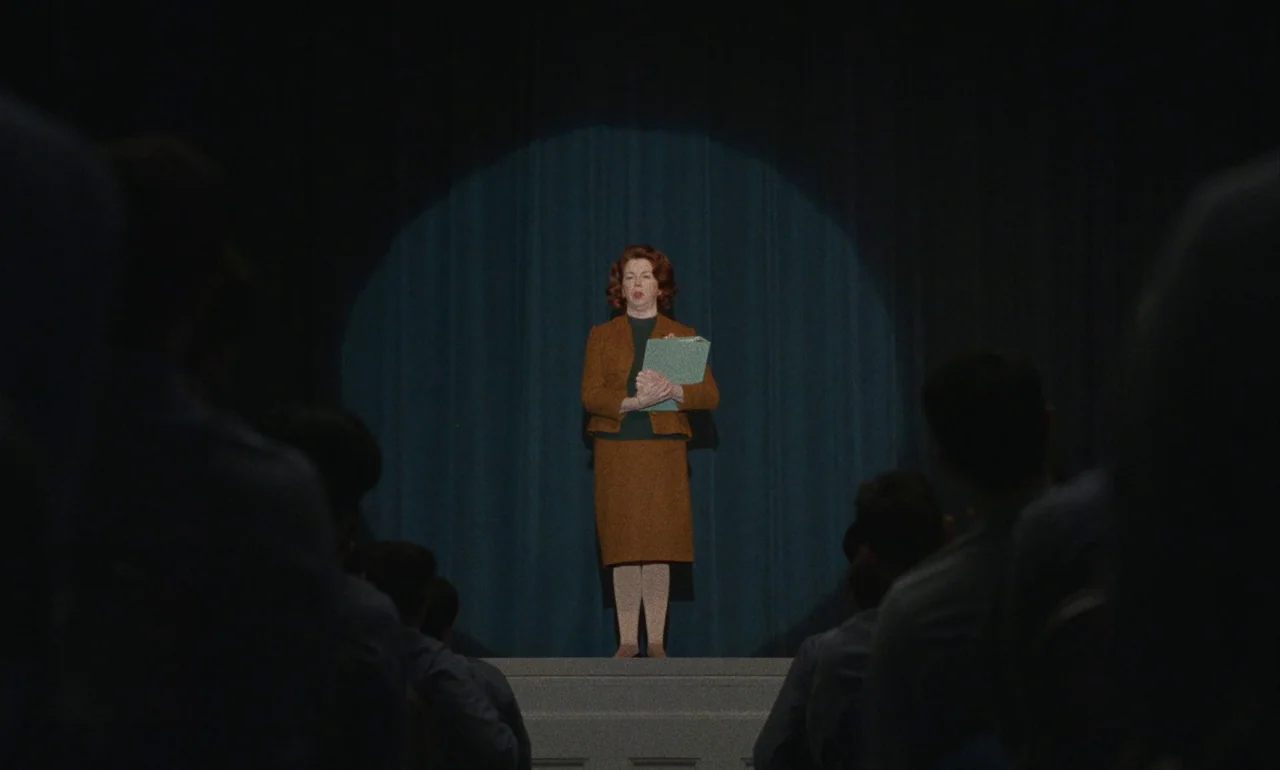 Eileen (2023) movie still: fullbody — An auditorium features a dark blue curtain backdrop and a central raised wooden platform. A middle-aged woman…