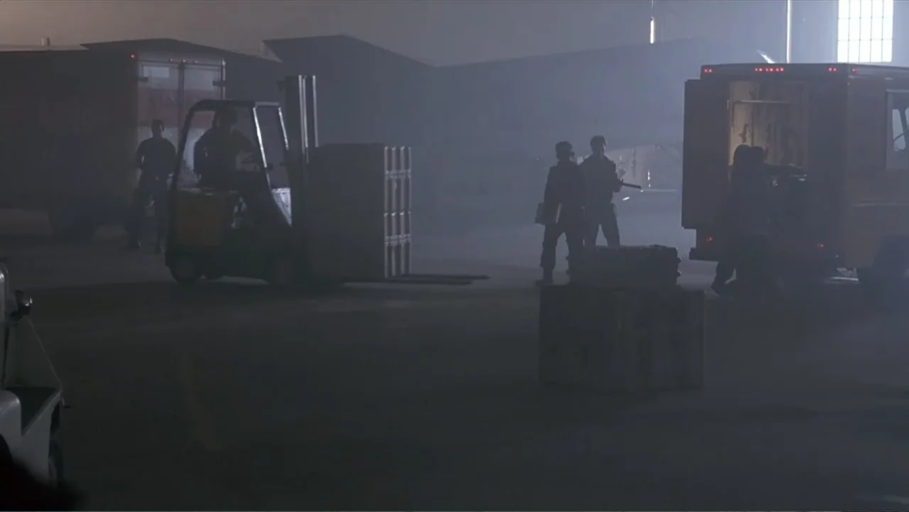 Crisis Point (2012) movie still: wide — A dark, misty industrial loading dock at night features parked trucks and large crates. Several workers in…
