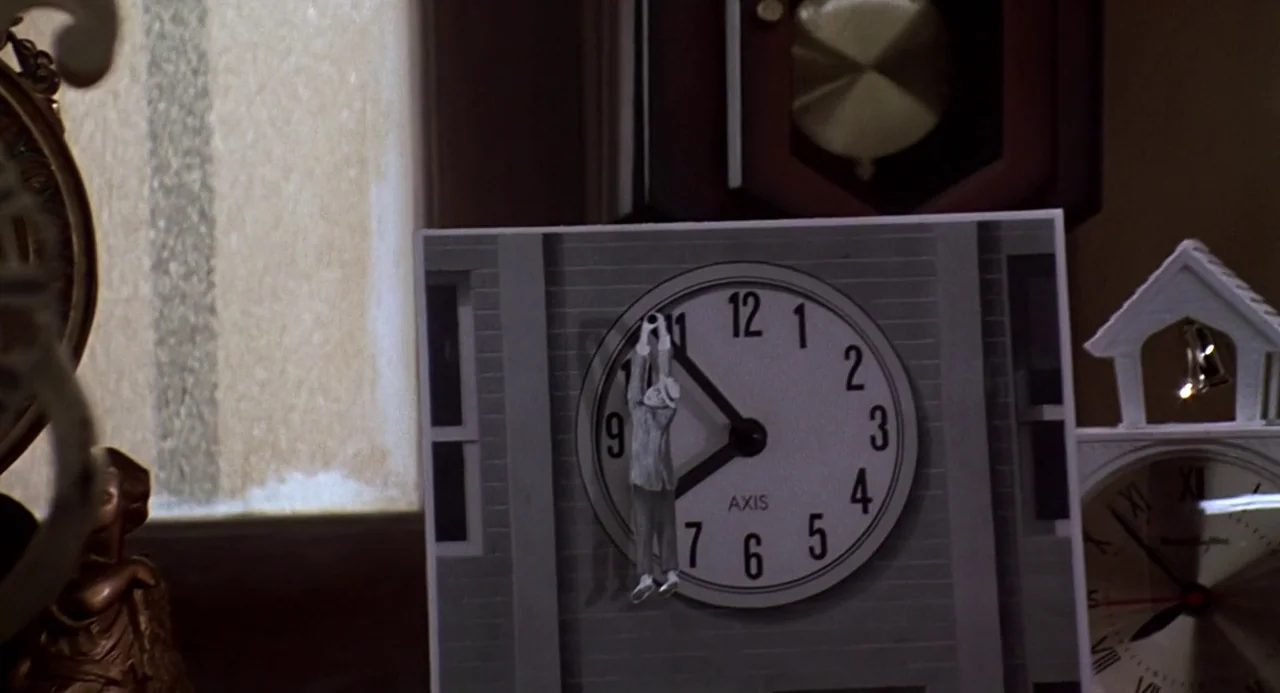 Back to the Future (1985) movie still: closeup — A miniature paper figure hangs from the minute hand of a stationary desk clock. Other clocks and…