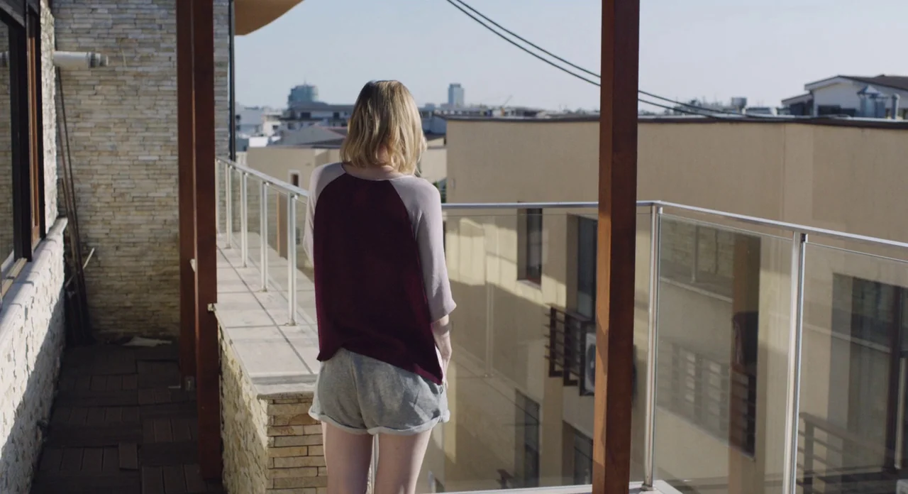 Toni Erdmann (2016) movie still: fullbody — An outdoor balcony with stone walls and a glass railing overlooks urban buildings. A woman stands with…