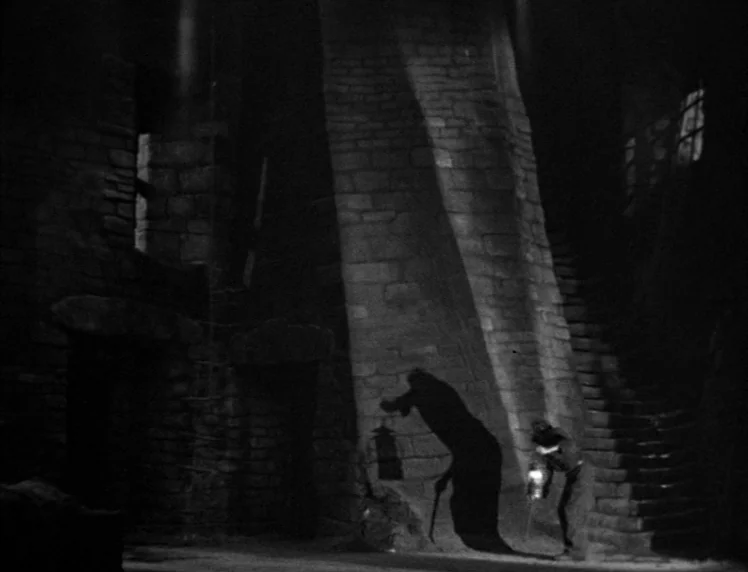 Frankenstein movie still: wide — A cavernous stone chamber features heavy masonry and a staircase built into the wall. A solitary figure walks…