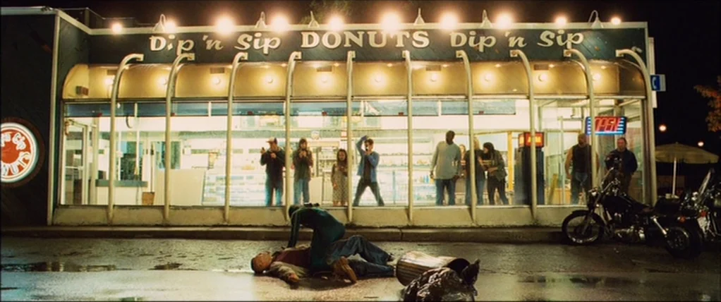 Kick Ass (2010) movie still: wide — A well-lit donut shop facade with glass windows and a line of spectators inside. A figure in a costume straddles…