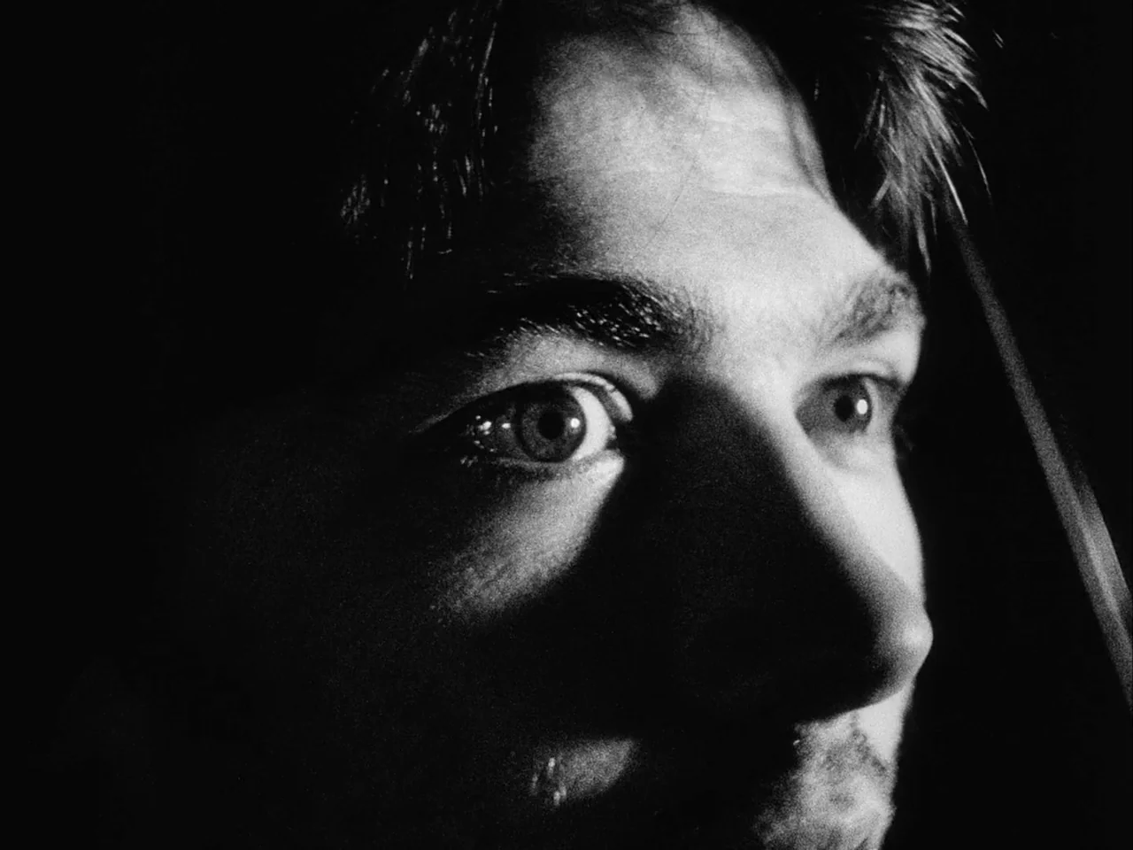 Mala Noche (1986) movie still: closeup — A man's face is partially obscured by darkness in a monochromatic setting. He gazes off-camera with a…