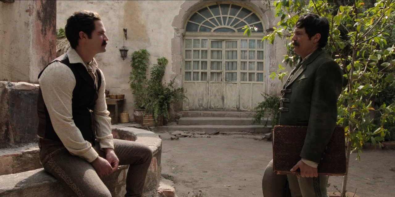 Pedro Páramo (2024) movie still: fullbody — A rustic stone-walled courtyard features a large arched window with a white grid frame. Two men in…