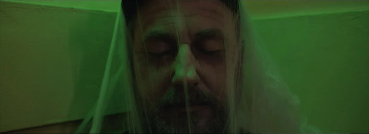 DannyBoy (2020) movie still: closeup — A man rests in a narrow corner lit by a distinct, vibrant green light. Thin translucent material hangs over…