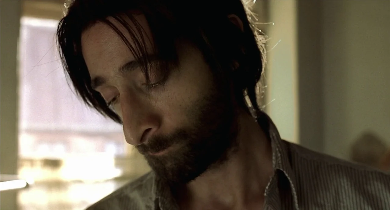 The Pianist (2002) movie still: closeup — A man stands indoors against a bright window source that backlights his hair. The man gazes downward while…