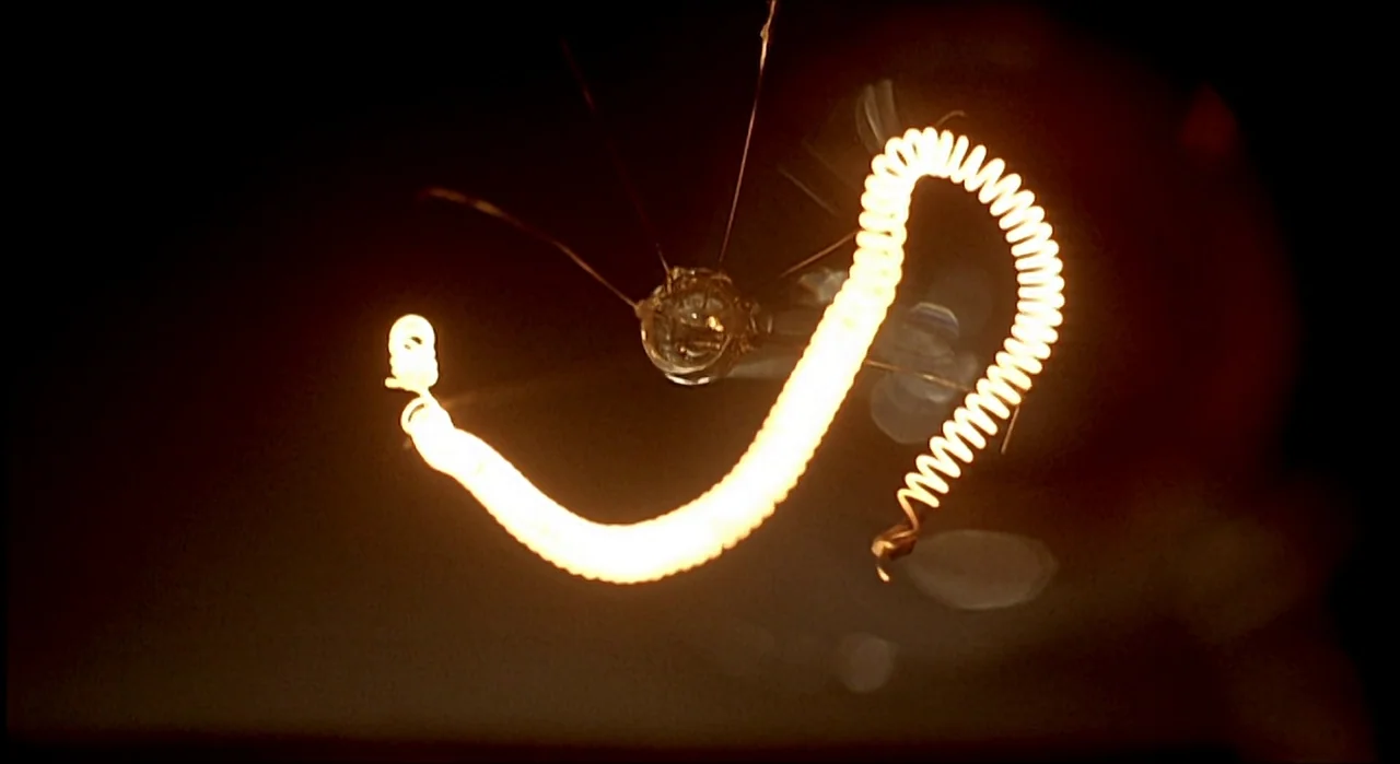Voice (2005) movie still: closeup — A close-up view of an incandescent light bulb filament glowing in the darkness. The fine metal coil illuminates…