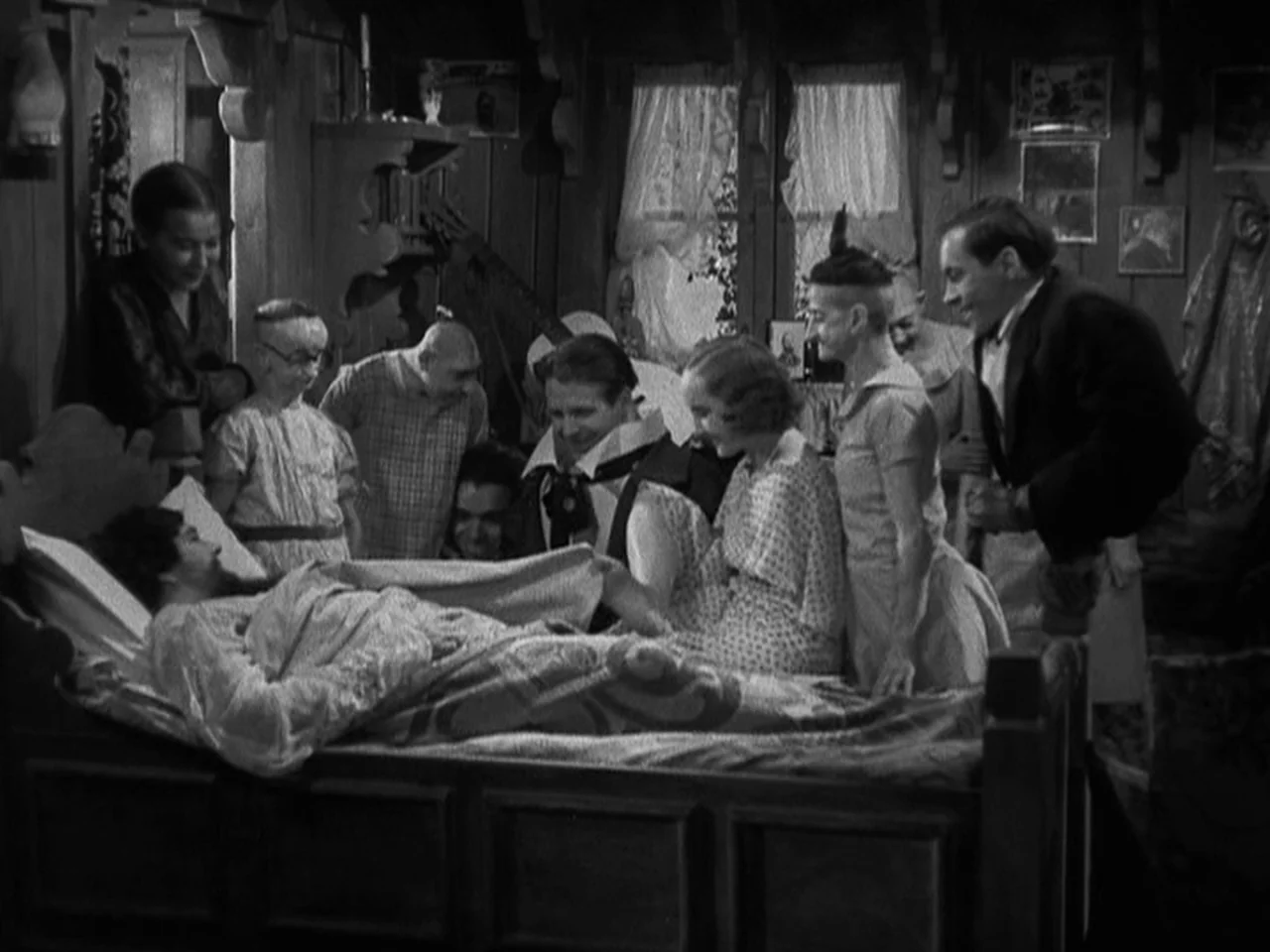 Freaks (1932) movie still: medium — A wooden cabin-style bedroom filled with a diverse group of people gathering around a bed. The figures stand…