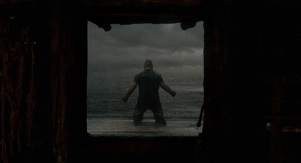 Noah (2014) movie still: wide — A dark wooden frame looks out onto a turbulent sea under a storm cloud. A muscular man kneels on the wet deck with…