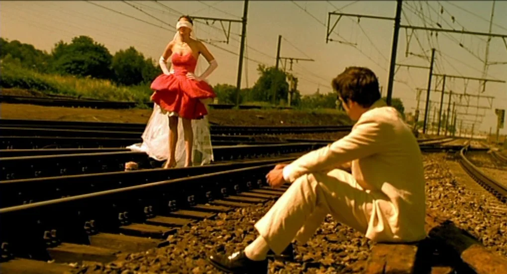Jeux d’Enfants (2003) movie still: fullbody — A set of railway tracks stretches into the distance under a bright yellow sky. A man sits on the rail…