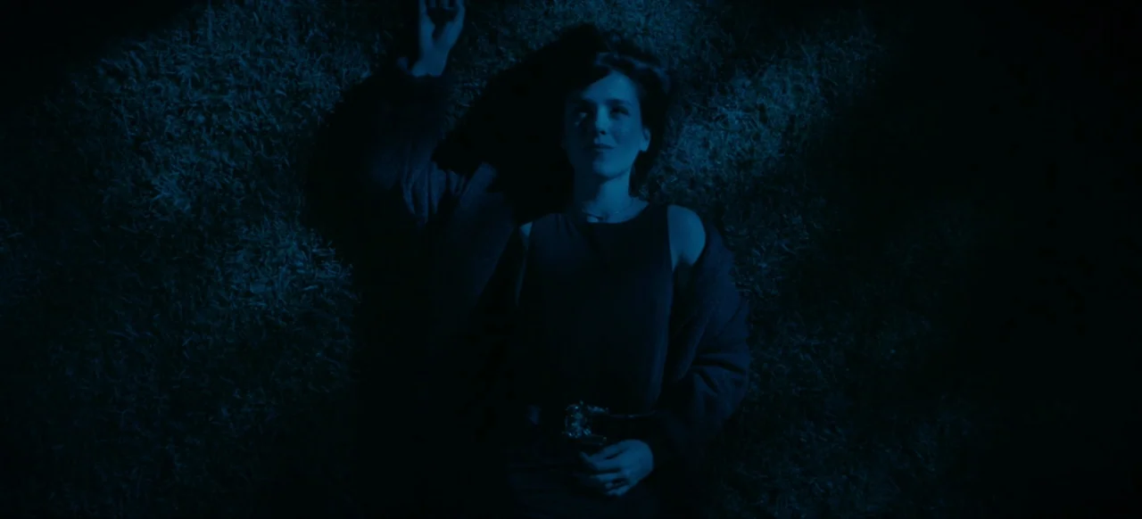 Housewife (2017) movie still: medium — A person lies flat on a dark patch of grass under night sky conditions. The subject is oriented looking up at…