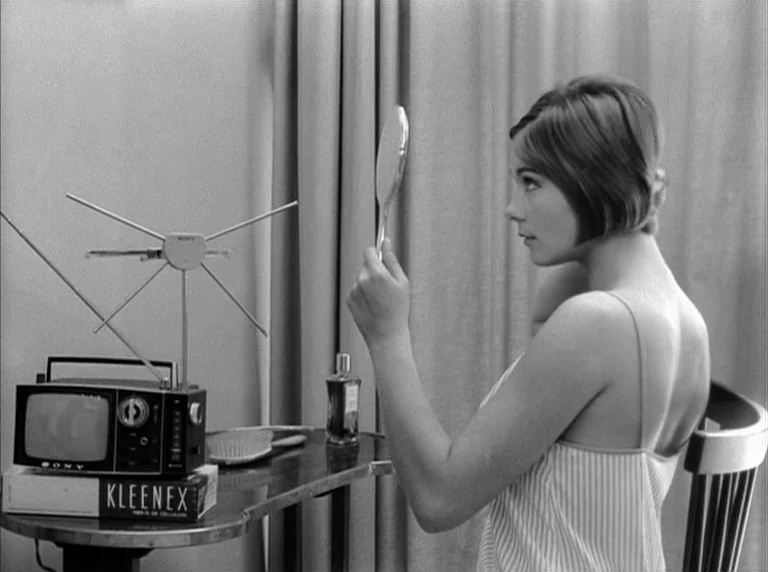 Une Femme Mariée (1964) movie still: medium — A woman sits at a vanity table with a television antenna and mirror. She looks into a small hand…