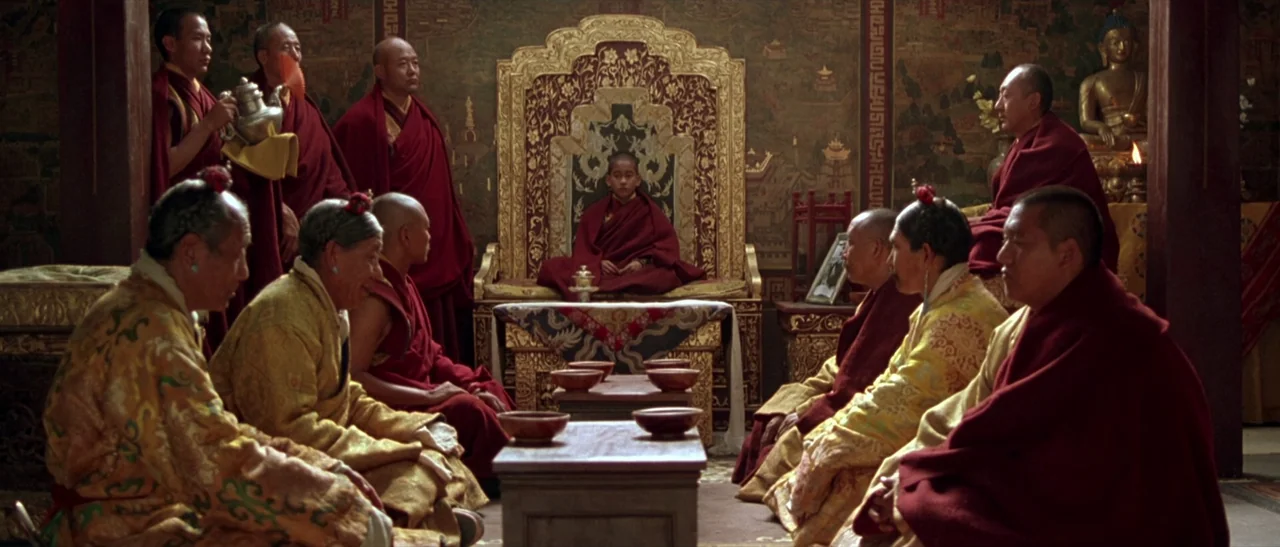 Kundun (1997) movie still: wide — An ornate interior temple features a central throne and numerous seated figures in traditional attire. Multiple…