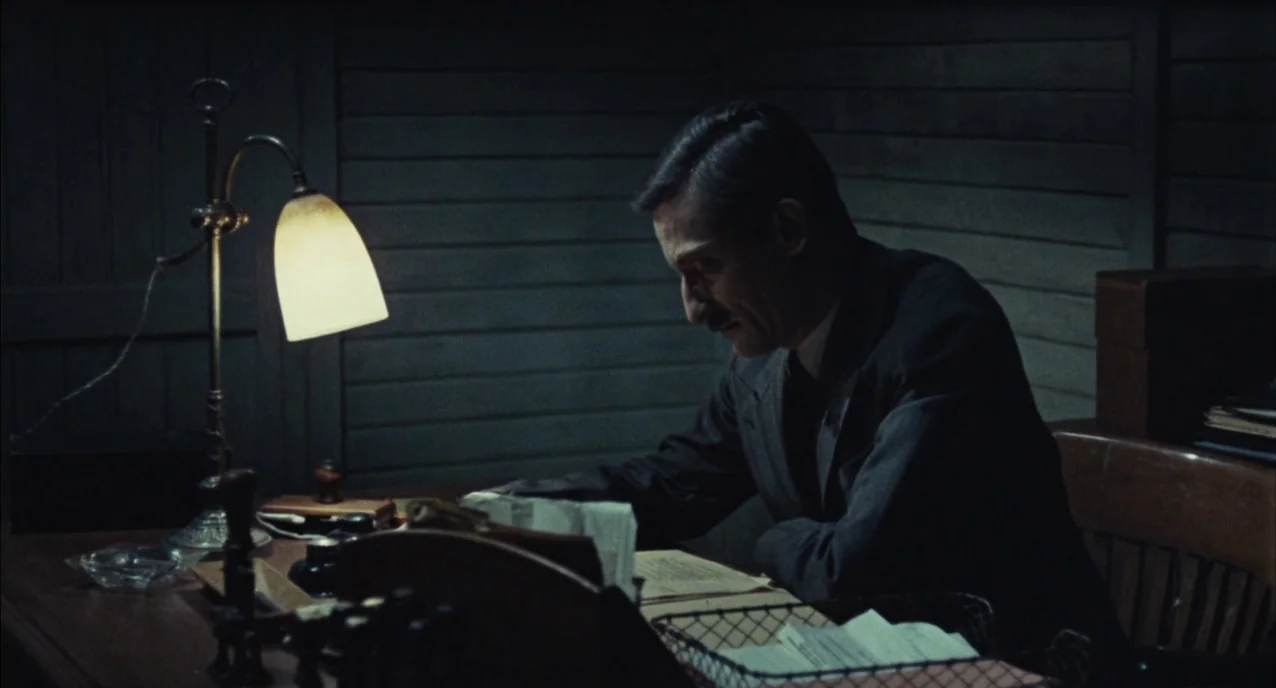 The Army of Shadows (1969) movie still: medium — An office with wooden walls and a cluttered desk containing documents and a lamp. A man sits at the…