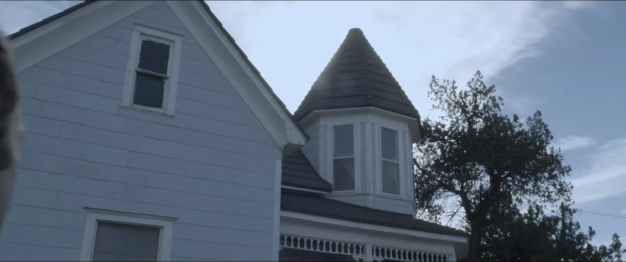 Amityville Cult (2021) movie still: establishing — A light blue wooden house with a turret stands against a bright sky. The building features dark…