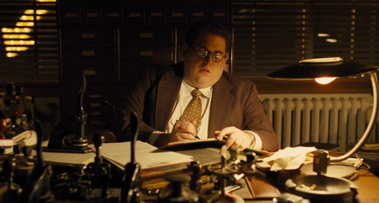 Hail, Caesar! (2016) movie still: medium — An dimly lit office features cluttered vintage rubber stamps and paperwork on a desk. A man wearing…