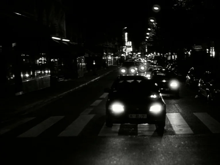 Eloge de L’amour (2001) movie still: establishing — A dark city street at night is illuminated by the bright headlights of oncoming cars. Painted…