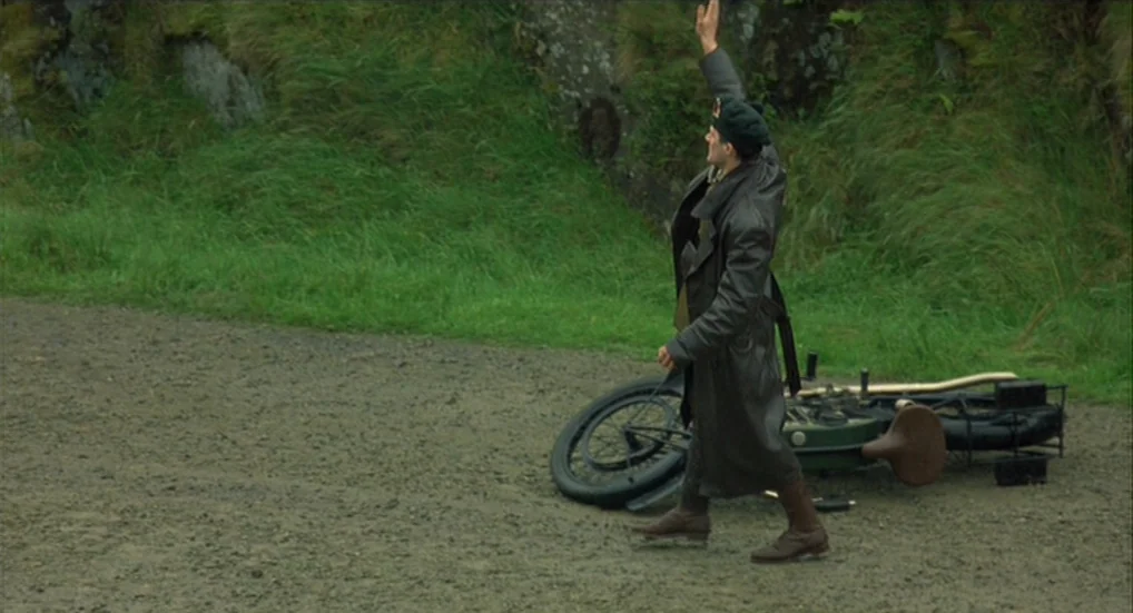 The Wind That Shakes the Barley (2006) movie still: fullbody — A gravel road sits at the base of a grassy, moss-covered stone embankment. A man in a…