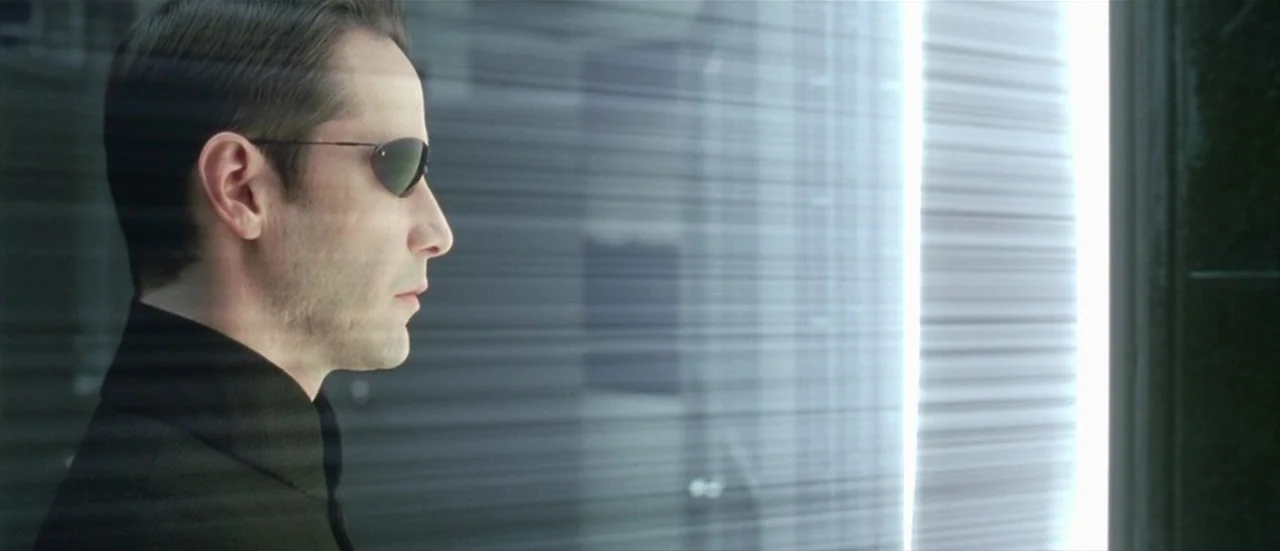 The Matrix Reloaded (2003) movie still: closeup — A man stands in an interior hallway with light filtering through horizontal blinds. He is…