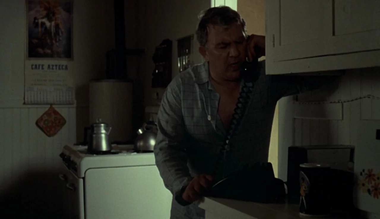 Fat City (1972) movie still: medium — A dimly lit 1970s interior with vintage appliances and a wall calendar. A middle-aged man stands at the…