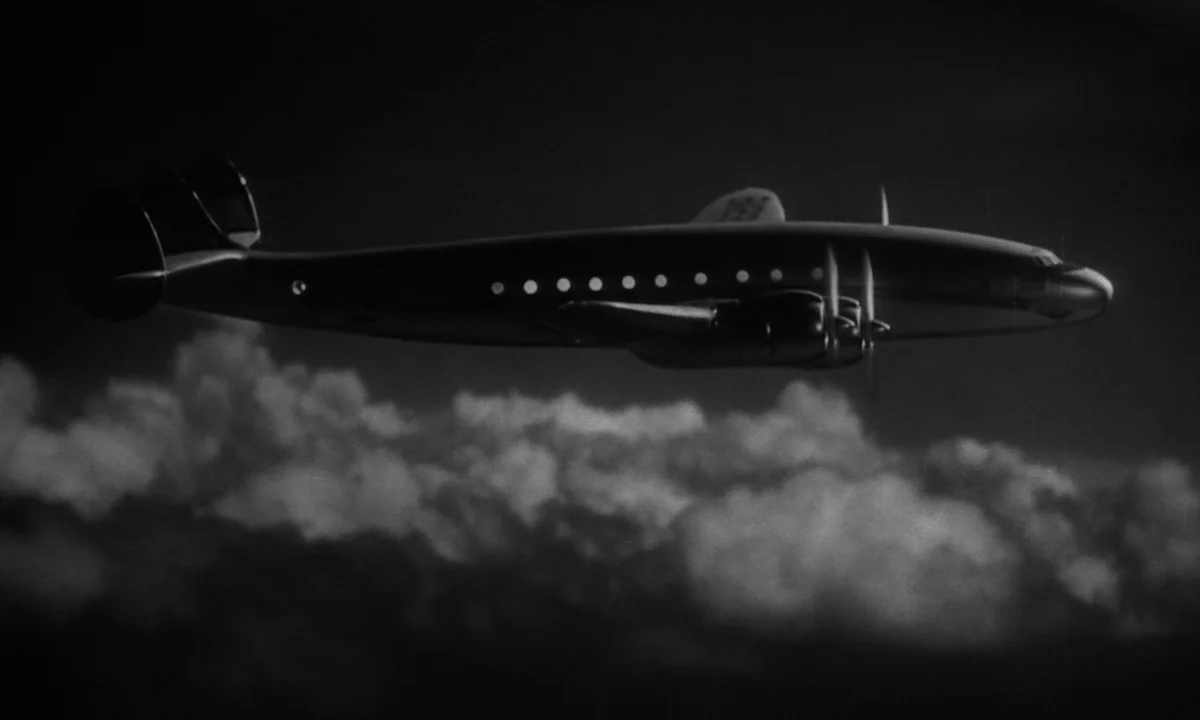 Night Of The Demon (1957) movie still: wide — A silver multi-engine airplane cruises through the night sky above a dense cloud layer. The dark…