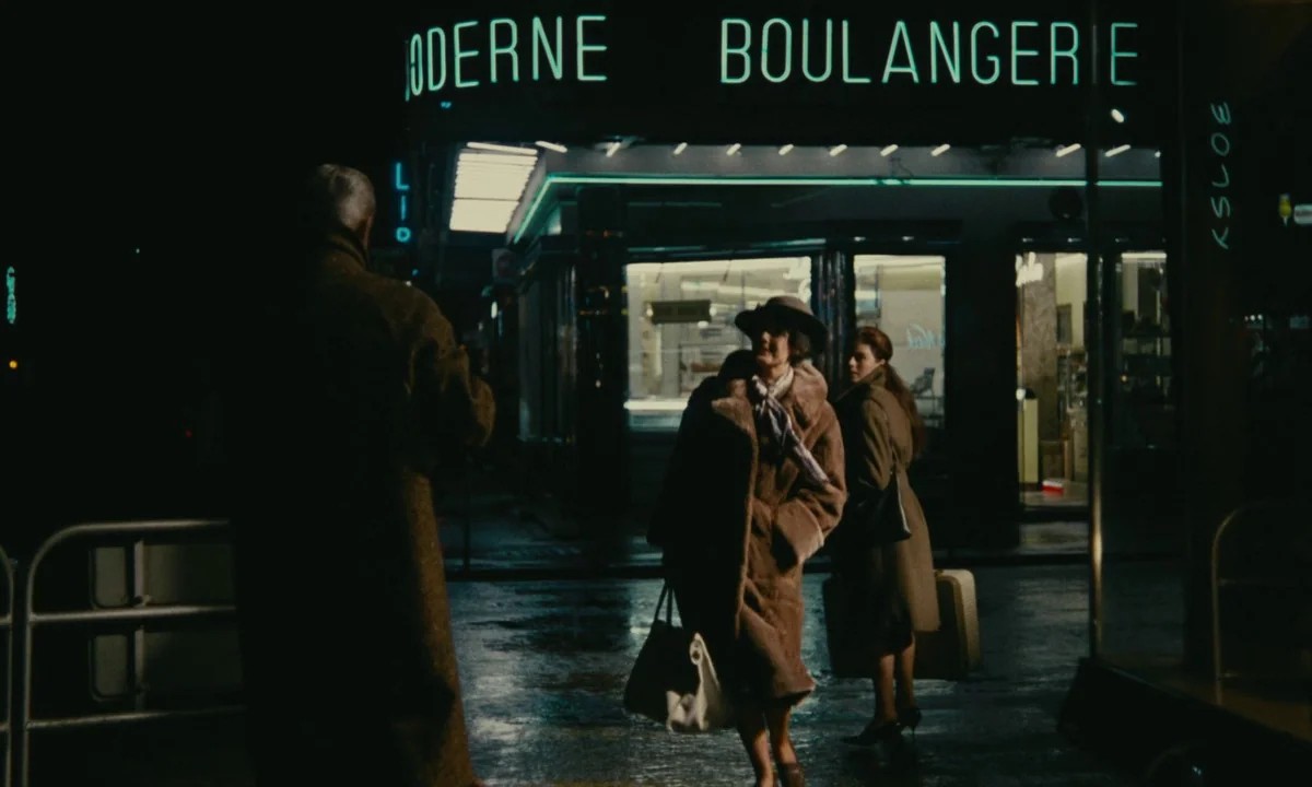Muriel (1963) movie still: wide — A dark, wet city street at night in front of a boulangerie with neon signage. A man in a coat watches two women…