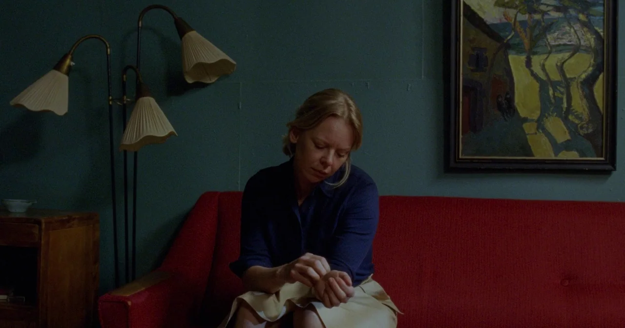 Fallen Leaves (2023) movie still: medium — A room with dark teal walls features a bright red couch and framed landscape art. A woman in a blue shirt…