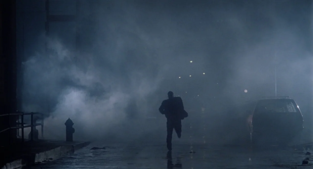 Def By Temptation (1990) movie still: wide — A dark city street at night filled with heavy rolling steam. A lone man runs away from the camera…