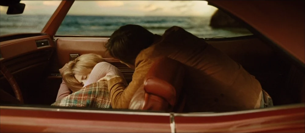 The Road (2009) movie still: medium — The interior of a vintage car with brown leather upholstery faces a coastal ocean sunset. A man leans across…