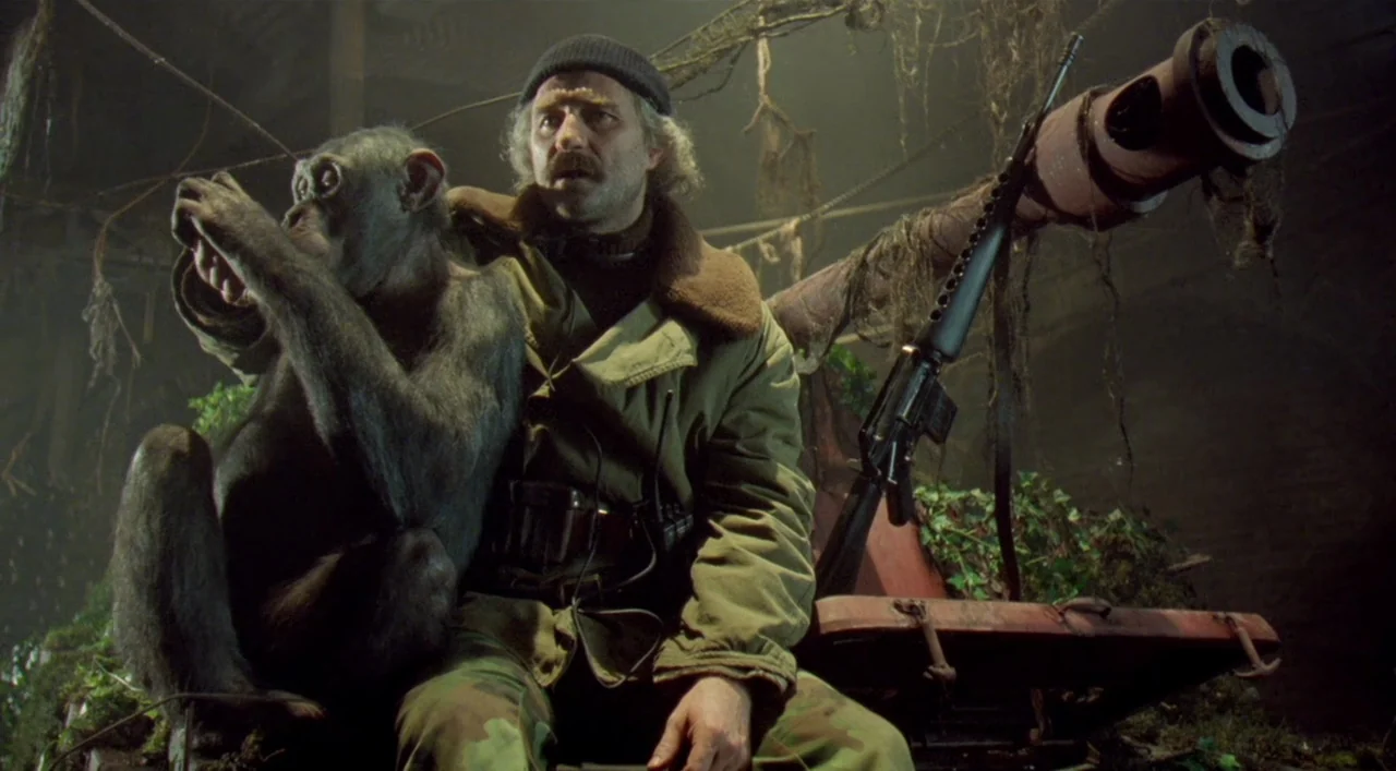 Underground (1995) movie still: medium — A man sits in an overgrown, dark, cavernous space alongside a chimpanzee. A large piece of rusted machinery…