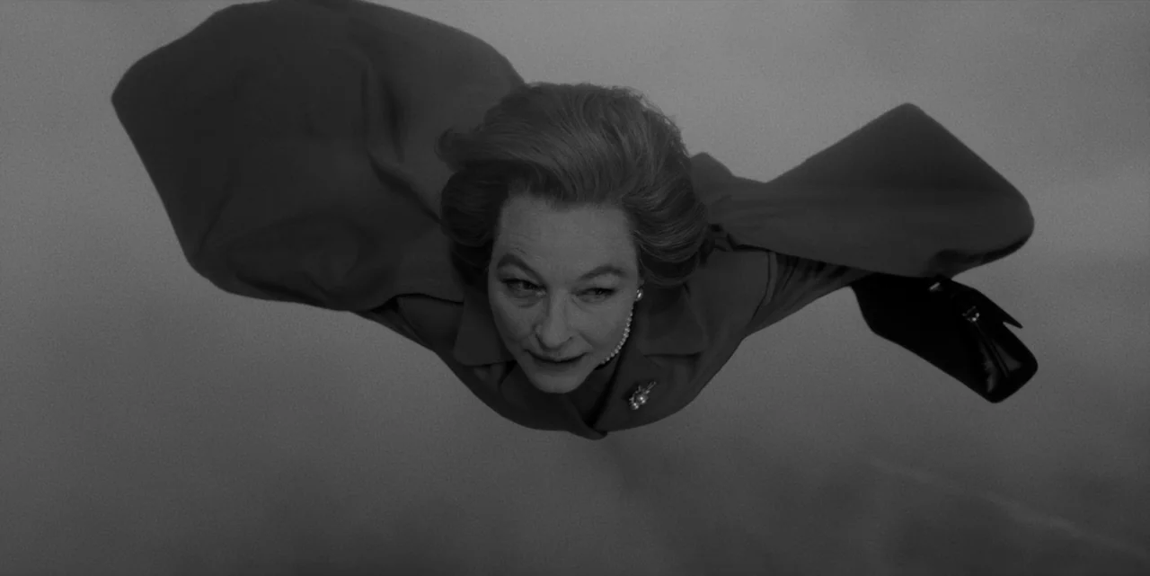 El Conde (2023) movie still: closeup — A woman drifts horizontally through an abstract, featureless gray void. The subject is oriented parallel to…