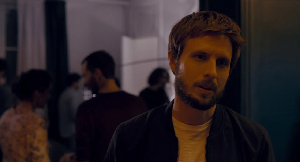 The Night Eats the World (2018) movie still: medium — An interior space at night with several people gathered in the soft-lit background. A man…