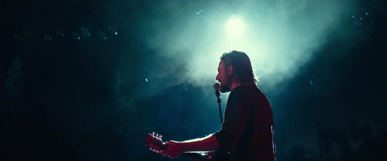 A Star Is Born (2018) movie still: medium — A dark outdoor stage filled with haze and illuminated by a bright rear light. A man in profile plays…