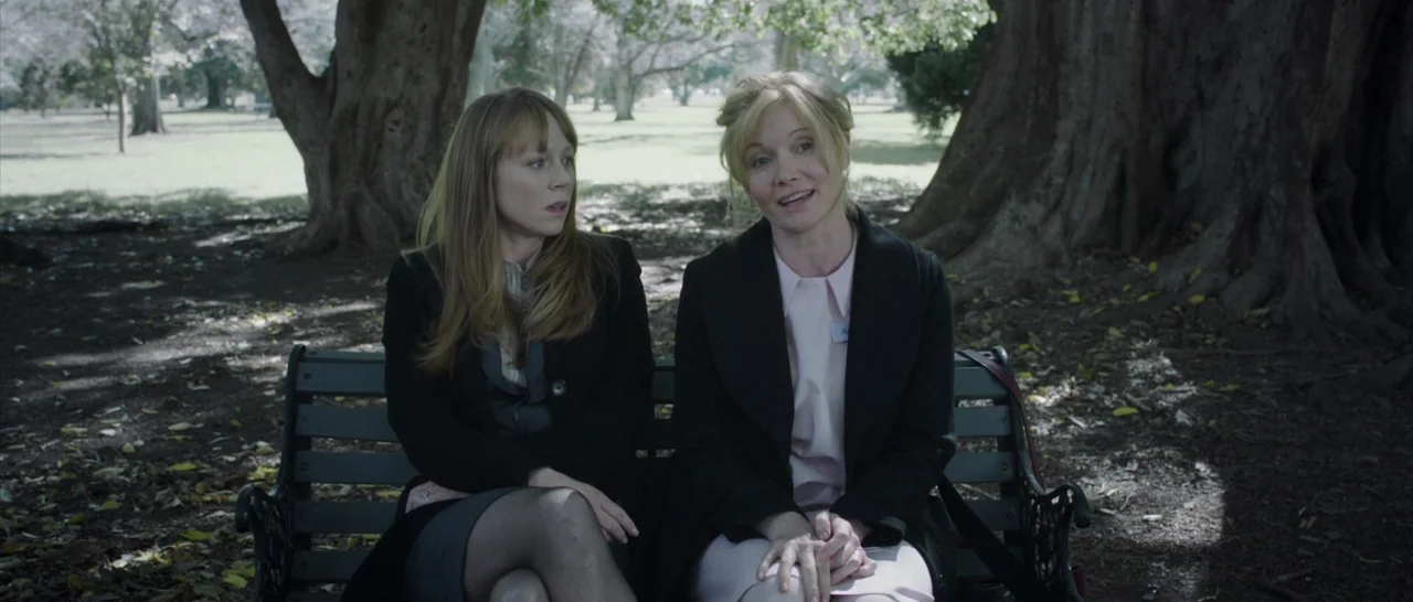 The Babadook (2014) movie still: medium — Two women occupy a metal bench beneath the dappled sunlight of a large tree. The background shows a public…