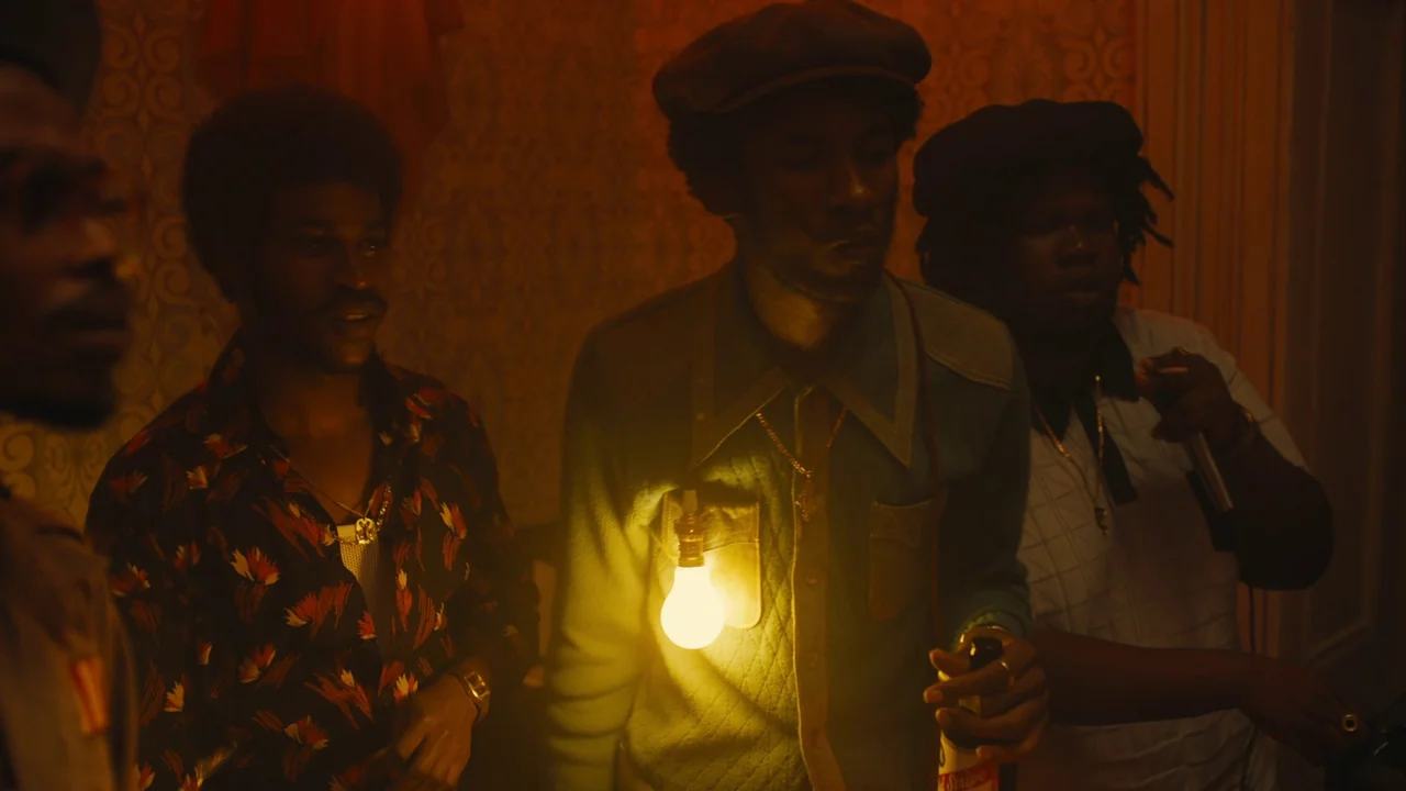 Lover’s Rock (2020) movie still: medium — A crowded, dimly lit indoor space with textured wallpaper and muted brown tones. Three men in…