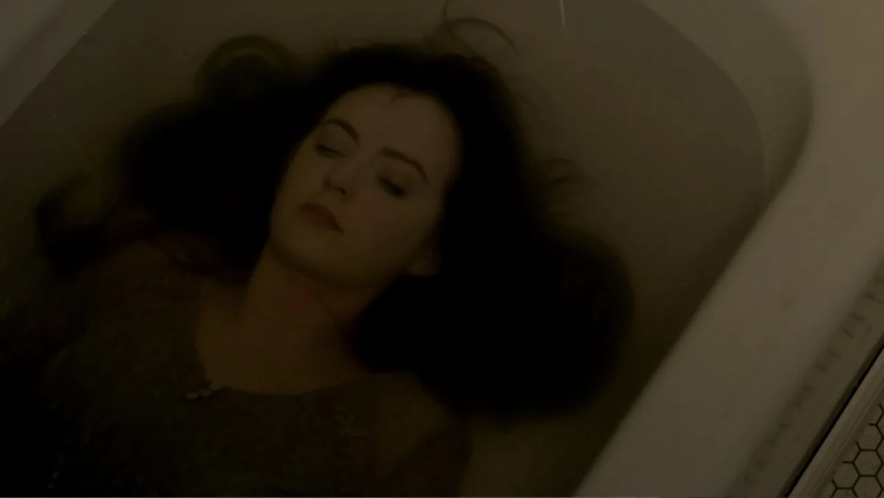Totem (2022) movie still: medium — A dark, interior bathroom scene with a person lying in a plain white porcelain tub. A woman with dark hair rests…