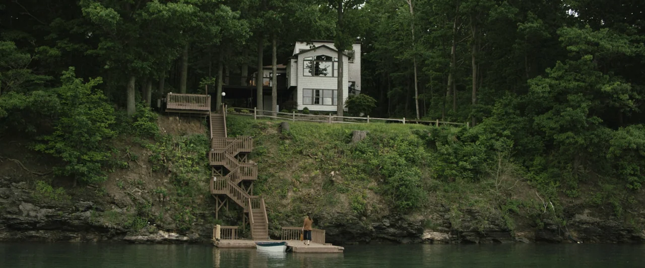 The Night House (2020) movie still: wide — A secluded white two-story house sits atop a grassy embankment overlooking a lake. A woman stands alone…