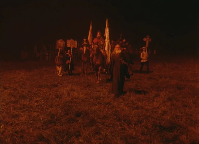 Russian Symphony (1994) movie still: wide — A group of people dressed in medieval garments walk across a dark, grassy field. They carry banners and…