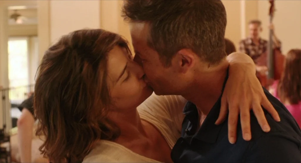 Results (2015) movie still: closeup — An interior space with a beige wall backdrop and soft natural light. A man and woman are embracing and kissing…