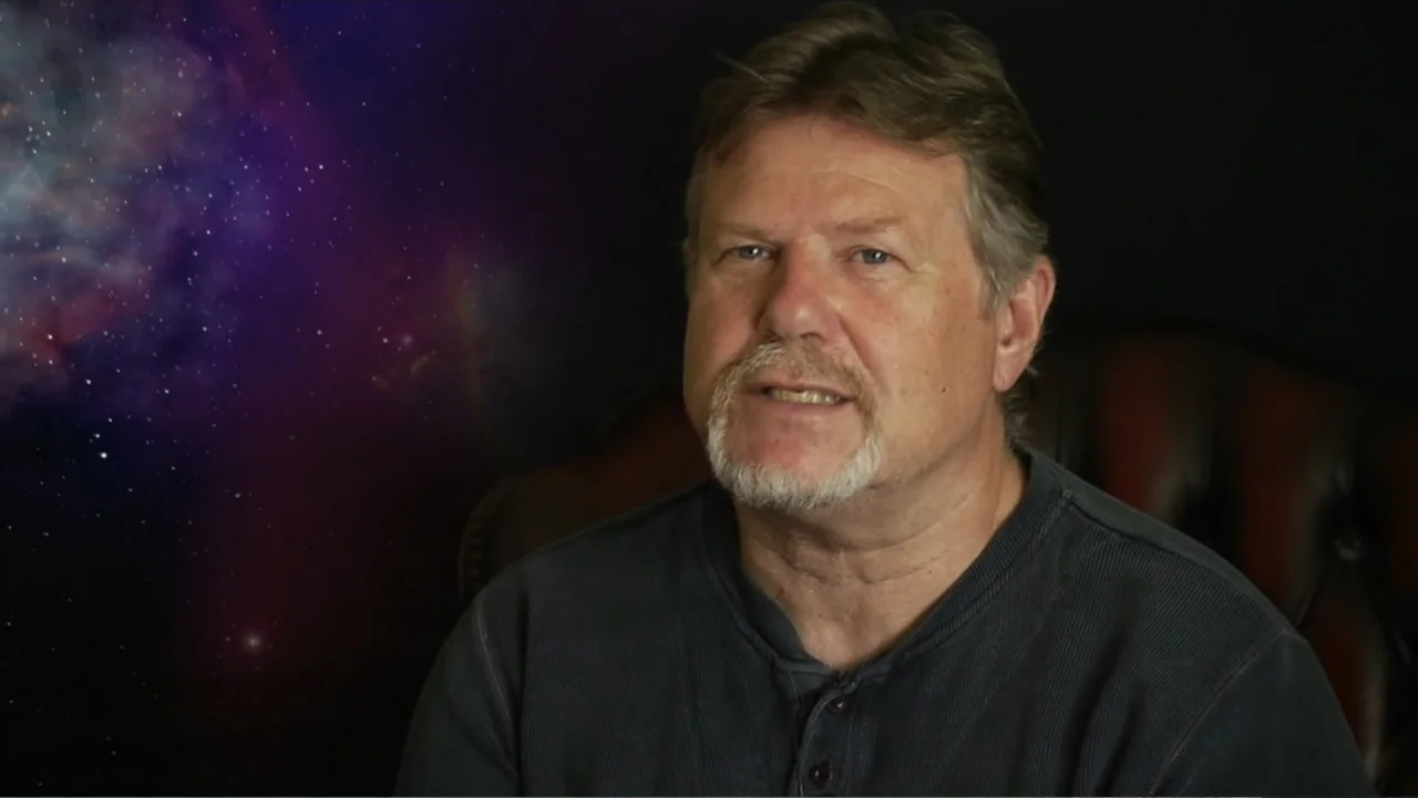 UFO Conspiracies: The Hidden Truth (2020) movie still: medium — A man sits in front of a dark backdrop featuring a projected image of space. The man…
