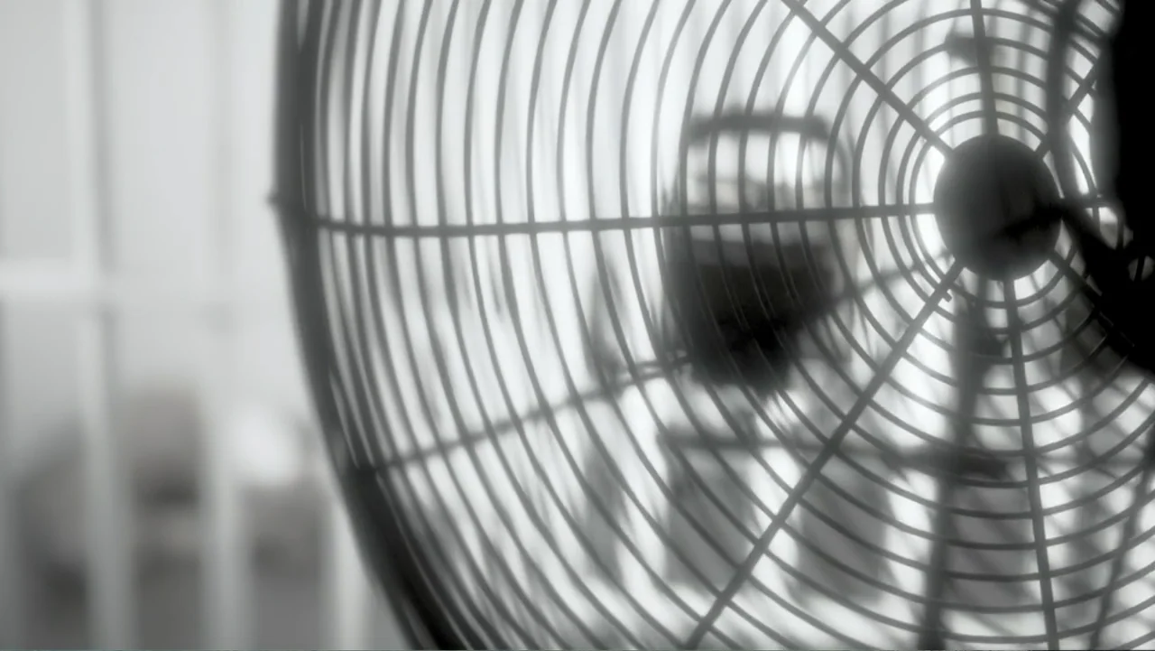 The Forever Prisoner (2021) movie still: closeup — An interior space contains a metallic electric fan in the foreground. The blades of the fan are…