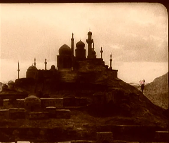 Destiny (1921) movie still: establishing — A miniature desert city model sits atop a rocky hill under a cloudy sky. The structure features multiple…
