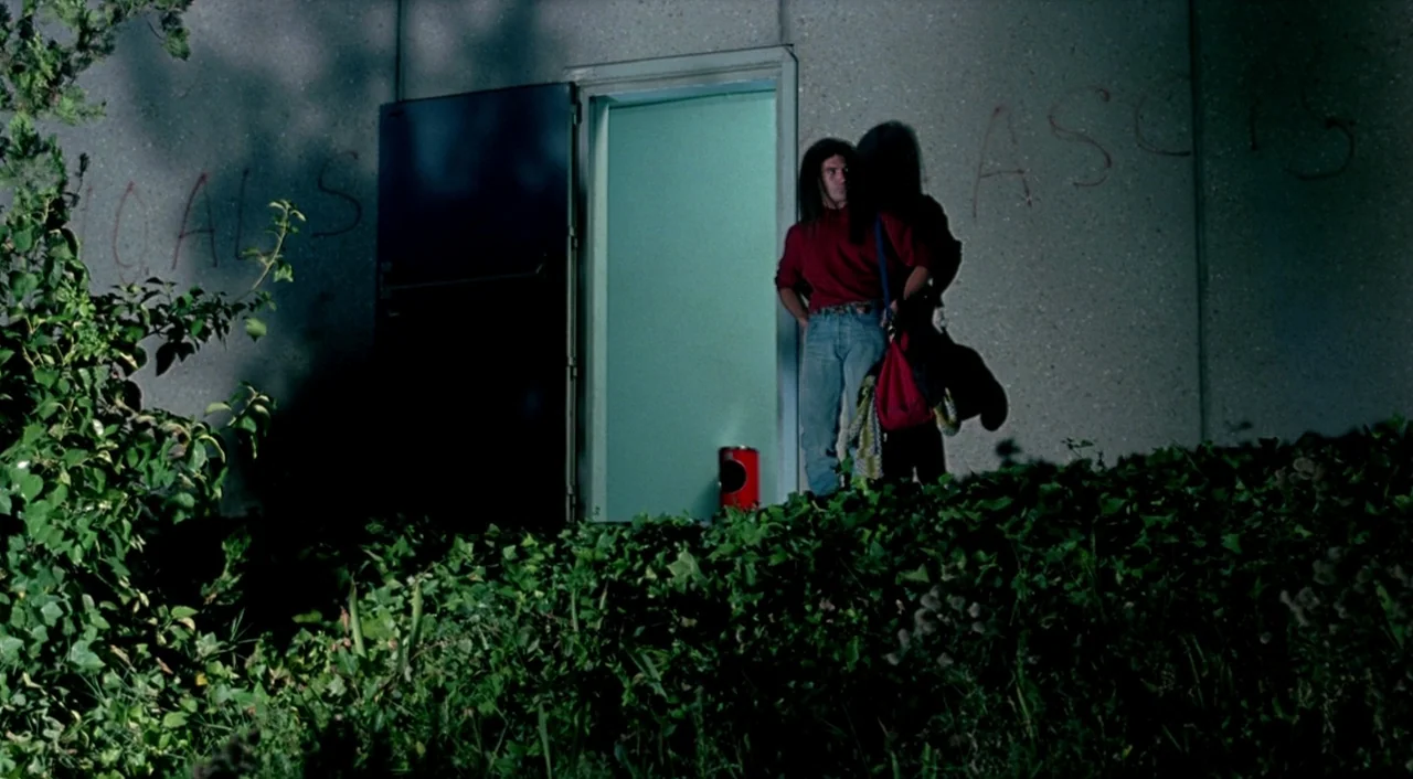 Tie Me Up! Tie Me Down! (1989) movie still: wide — A light-colored building wall has red graffiti lettering around an open dark doorway. A man…