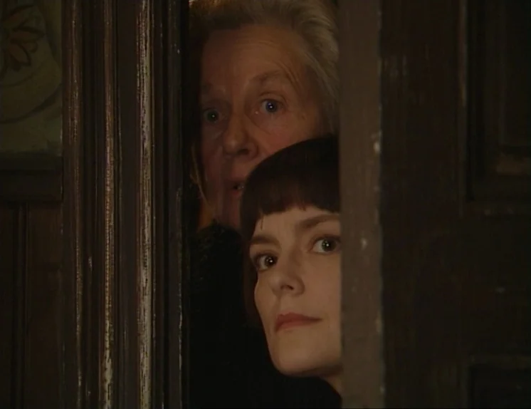 In The Presence of a Clown (1997) movie still: closeup — Two women peer through a vertical gap in a dark wooden door frame. The younger woman is in…