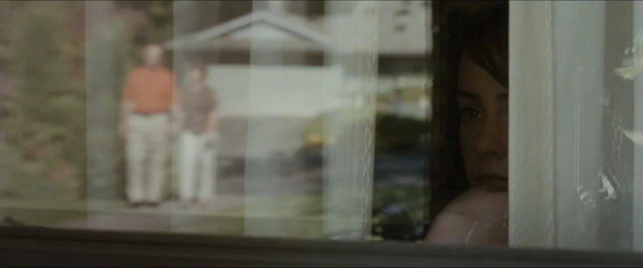 Into the Wild (2007) movie still: closeup — A woman looks through a glass window from the interior toward an exterior suburban house. Two blurry…