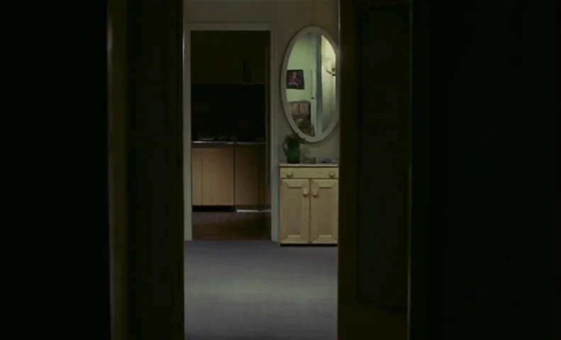 No End (1985) movie still: wide — A dark hallway leads into an illuminated room featuring a light-wood cabinet and mirror. The interior space…