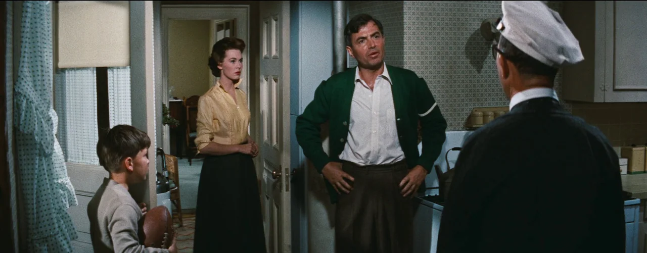 Bigger Than Life (1956) movie still: medium — A 1950s kitchen with patterned wallpaper and plain wooden cabinets. A group of four people gather in…