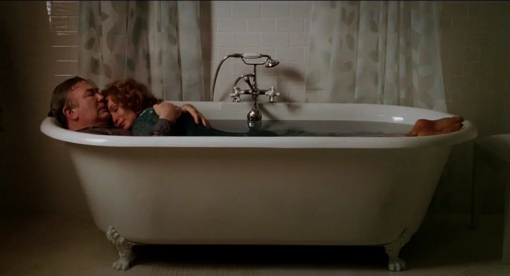Big Fish (2003) movie still: wide — A dark, tiled room features a prominent white freestanding clawfoot bathtub. An older couple in dark clothing…