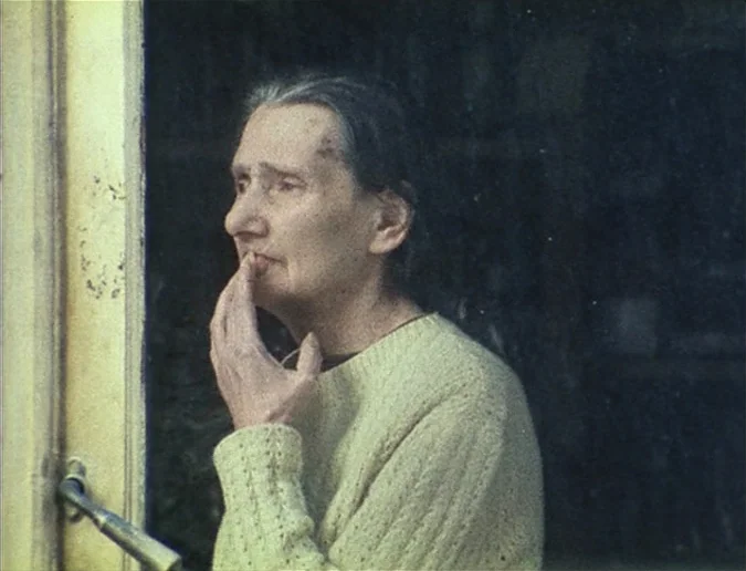 Daguerréotypes (1976) movie still: medium — An elderly woman stands indoors near a window frame. She rests her chin on her hand while looking…